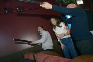 Three excited guests, a woman and two men, holding mock shotguns and laughing while participating in an interactive activity at the Nebula event