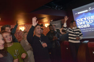 Group of enthusiastic attendees with drinks cheering and raising their hands in a dark cinema room at the Nebula Oxygen Influencers event