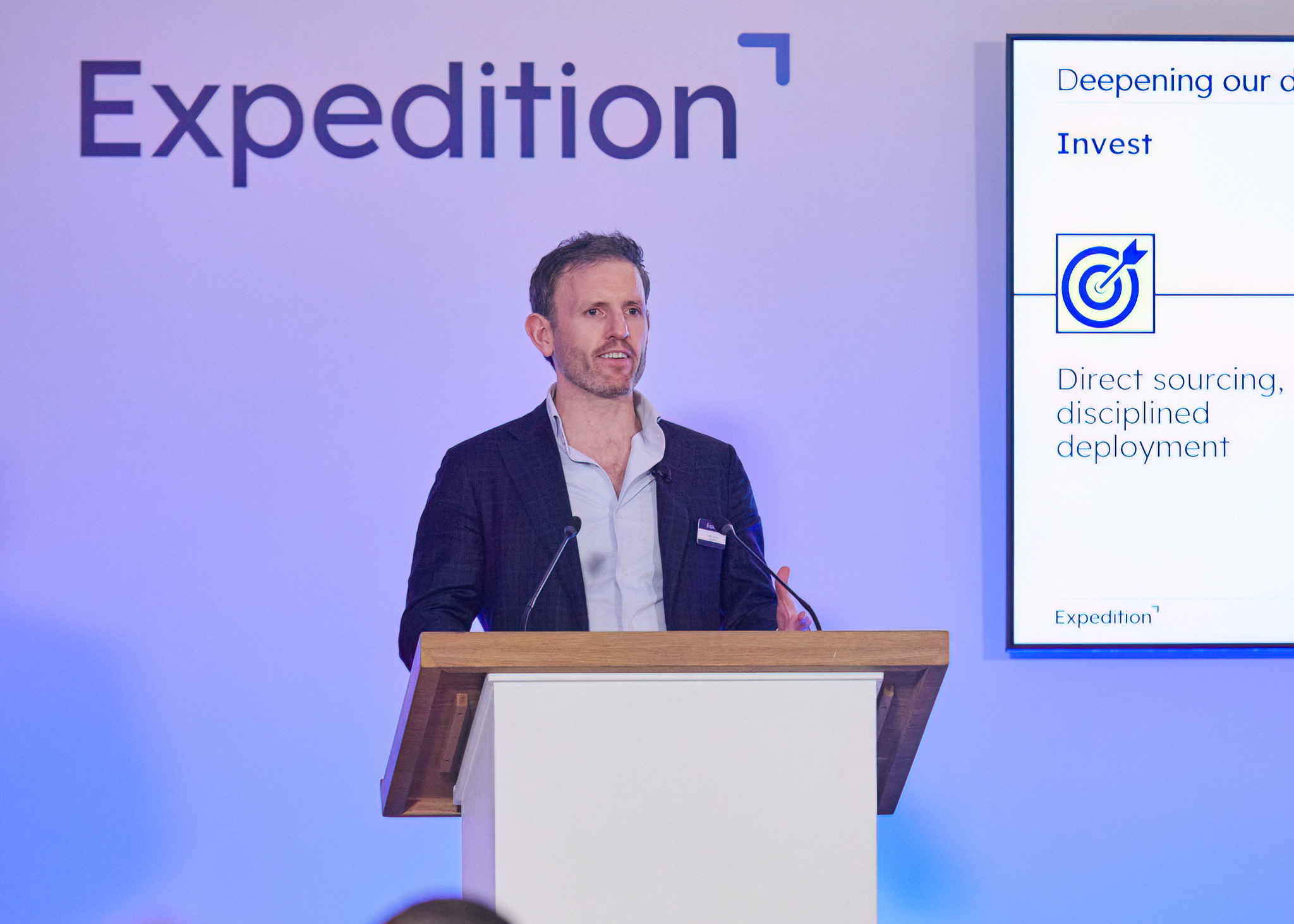 A speaker presenting at Expedition Capital's investor day, standing at a wooden podium in front of a large branded backdrop, with a slide visible on screen highlighting the firm's investment approach of direct sourcing and disciplined deployment.